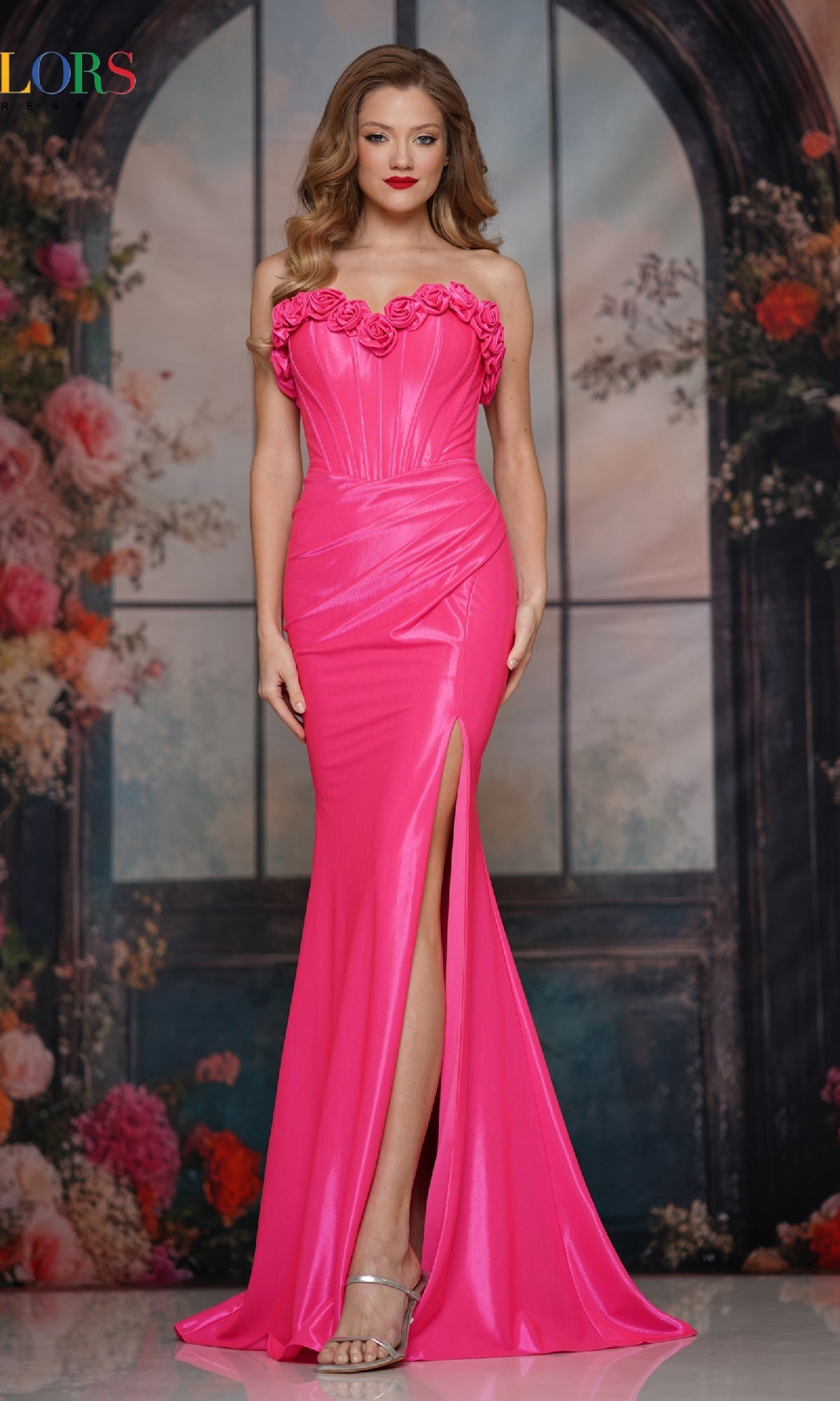 A woman stands in front of a floral and window backdrop wearing the Strapless Long Rosette Prom Gown: Colors Dress 3895, featuring a bright pink hue, sweetheart neckline, rosette detailing, and a thigh-high slit.